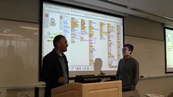 Two people standing in front of a large screen displaying a block-based programming interface.