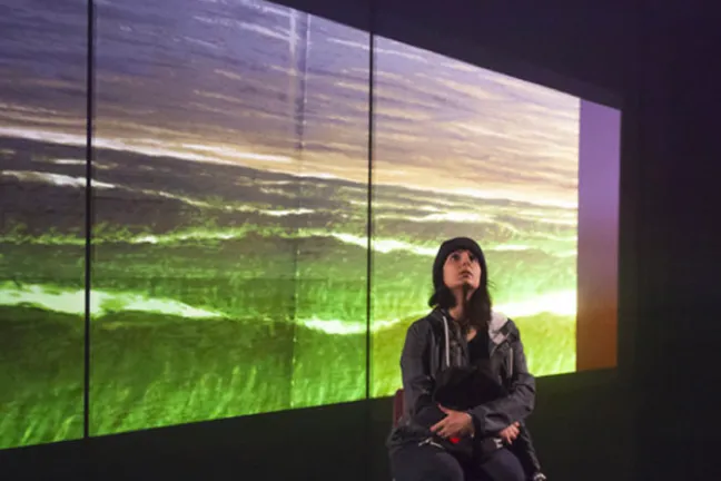 A person seated on a stage looking up, in front of seaside background.