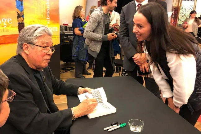 A person seated and signing a book for another individual.