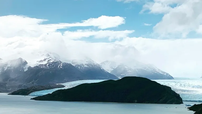 Glacier descending into a lake, backed by snow-covered mountains and a cloudy sky.