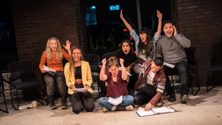 Cast of Towards Youth celebrating with their arms raised during a rehearsal